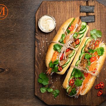 Two fresh Vietnamese banh mi sandwiches on a wooden board with herbs and chili.