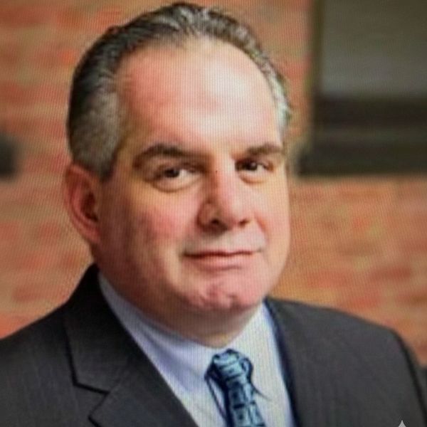 Professional middle-aged man in a suit and tie posing against a blurred brick background.