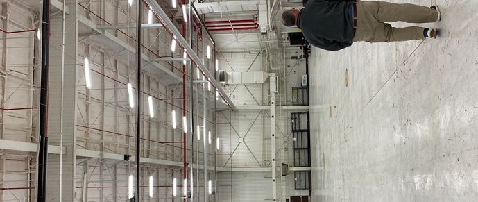 A person walks inside a large empty warehouse with high ceilings and bright lights.