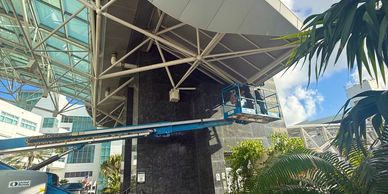 A worker on a mechanical lift performs maintenance on a building's exterior under a large canopy.