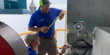 Two workers measuring near a security camera installation on an industrial site.