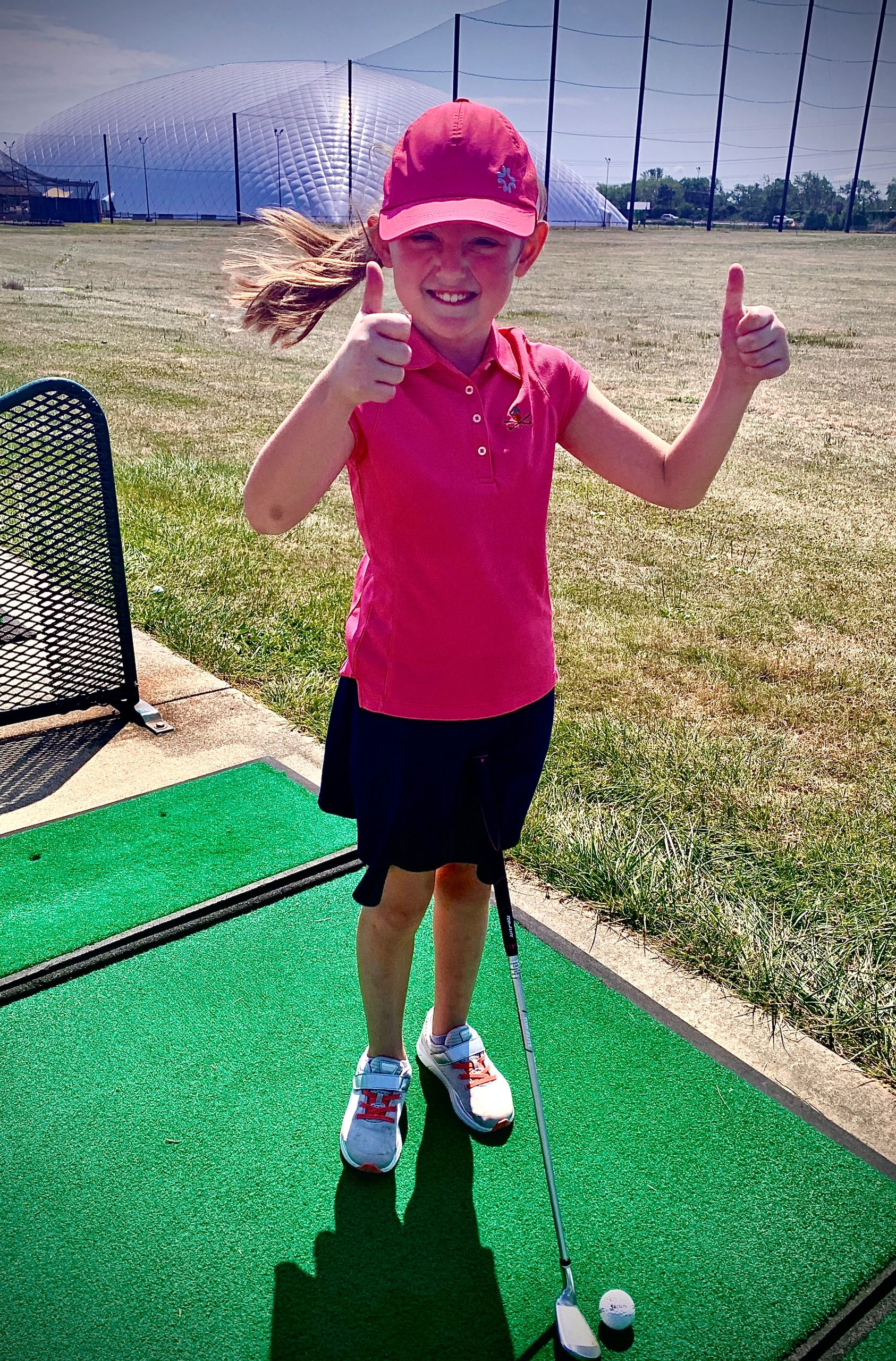 Young girl in pink golf outfit giving two thumbs up at a driving range.