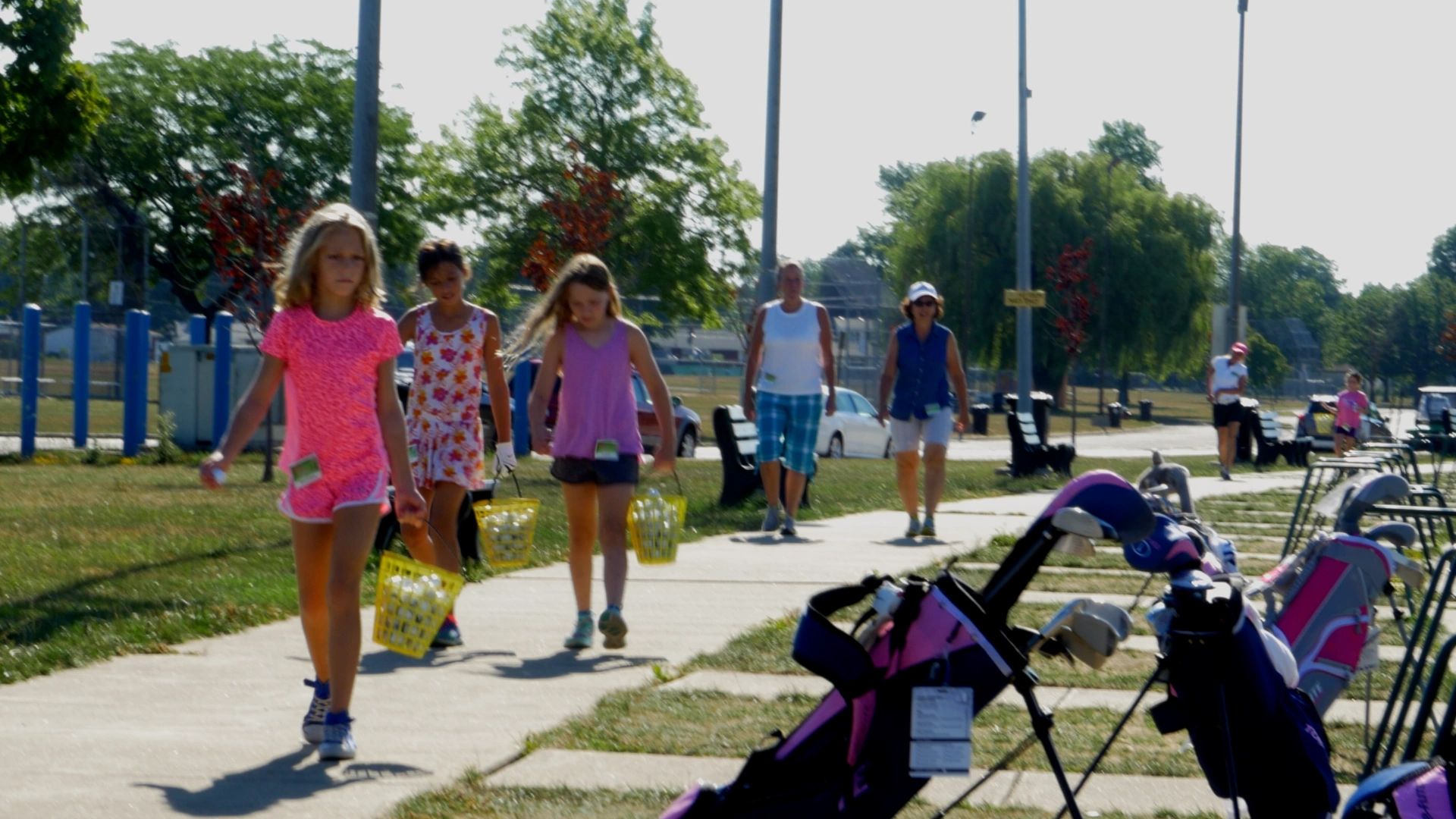 Children and adults walk on a path carrying baskets of golf balls at a golf practice range.