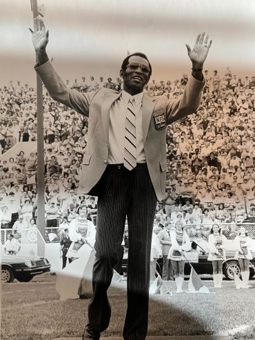 Man in NFL jacket waving to a large crowd at a stadium.