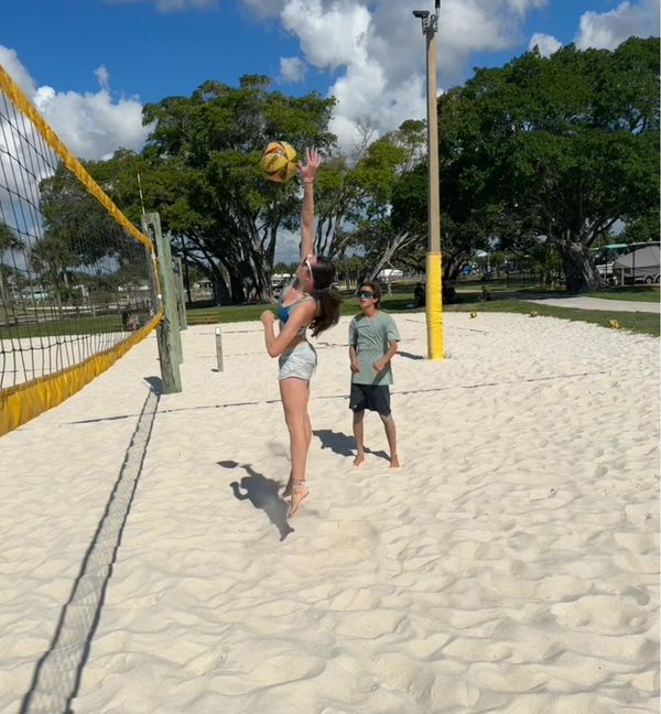 A girl jumps to hit a volleyball over the net on a sunny beach court.