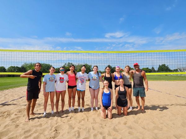 Group of young volleyball players posing on sand court under blue sky.