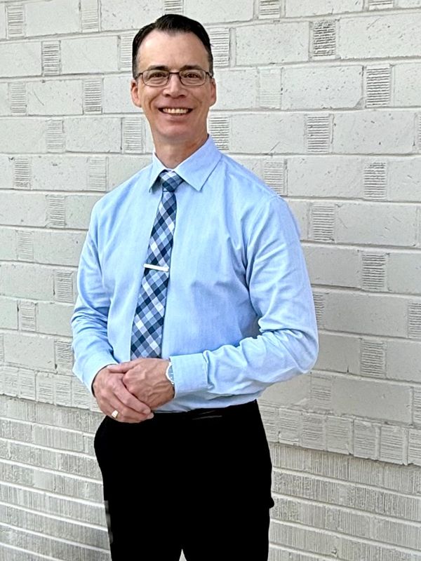 Man in a blue dress shirt and plaid tie standing against a white brick wall.