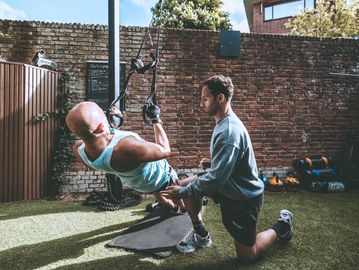 Trainer assists man doing suspension training outdoors.