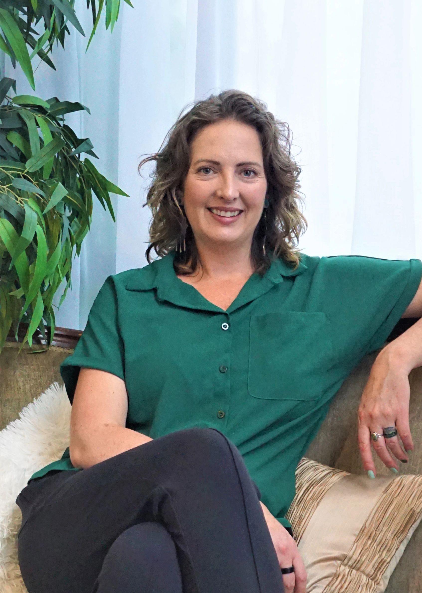 A woman in a green shirt smiles while sitting on a couch near a plant.