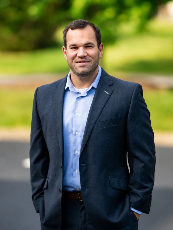 Man in a dark suit and light blue shirt standing outdoors.