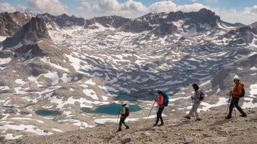 Four hikers traverse a rocky mountain ridge with snowy peaks and turquoise lakes below.