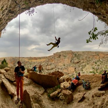 People rock climbing inside a large cave with cloudy sky.