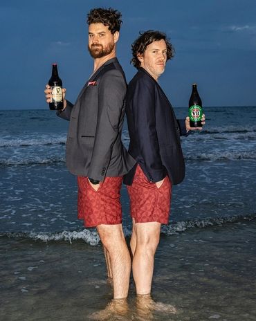 Two men in blazers and red shorts stand back-to-back in shallow water holding beer bottles.