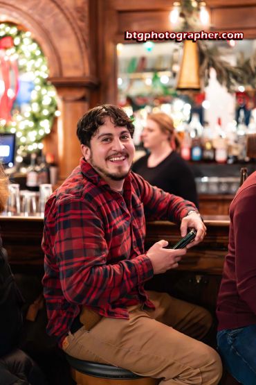 A smiling man in a red plaid shirt at a festive bar.