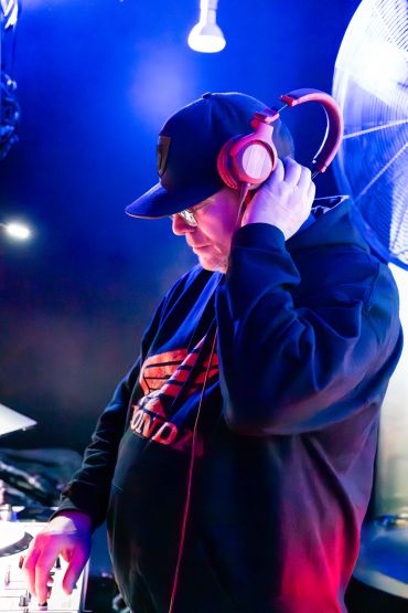 DJ wearing headphones and a cap, focused on mixing music in a dimly lit room.