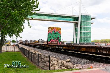 Graffiti-covered train car passing under a modern pedestrian bridge.