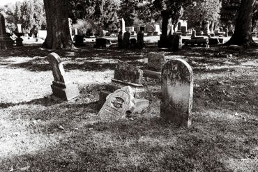 Old, weathered gravestones in a shaded cemetery surrounded by trees.