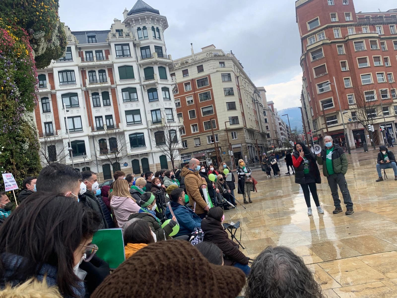 A crowd gathers in a city square for a public event with speakers and masks.
