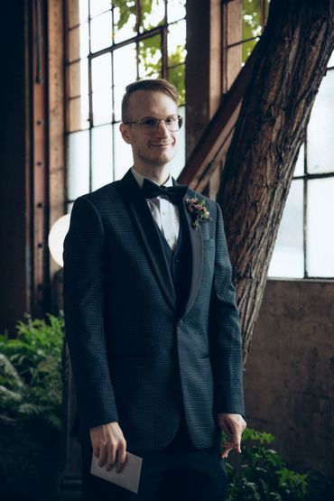 Man in tuxedo with boutonniere stands indoors near large windows.