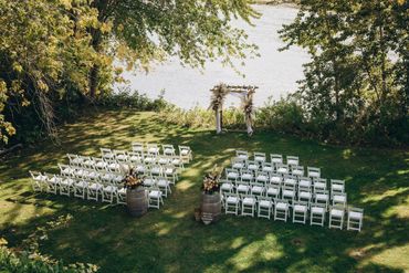 Outdoor wedding setup with white chairs and floral arch by the water.