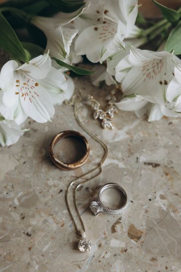 Wedding rings, a diamond necklace, and white flowers on a marble surface.