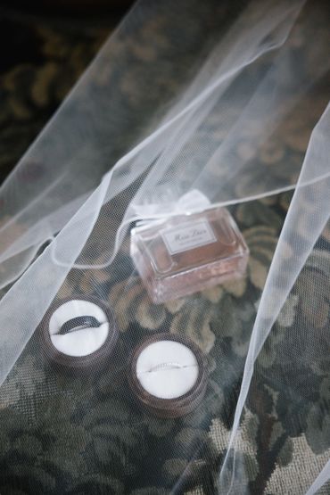 Wedding rings and perfume bottle arranged on a patterned cloth under sheer fabric.