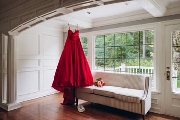 Elegant red dress hanging in a bright room with a bouquet and shoes nearby.