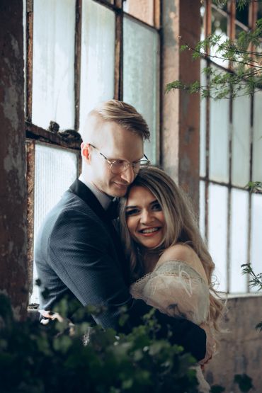 A couple embracing happily by large rustic windows with greenery around.