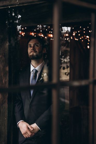 Man in a suit standing contemplatively indoors with soft string lights in the background.