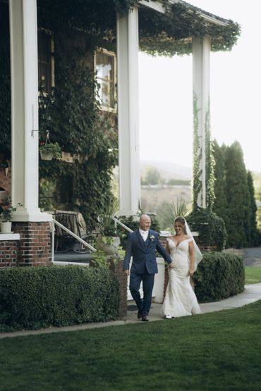Bride and groom walking hand in hand outside a vine-covered house.
