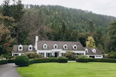 Large white house with manicured bushes and green lawn against forested hills.