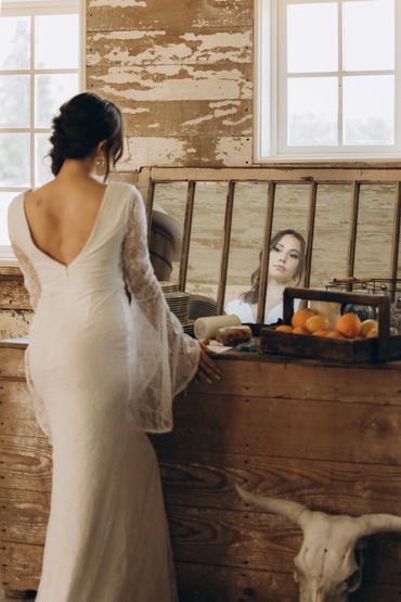 A woman in a white dress gazes at her reflection in a rustic room.