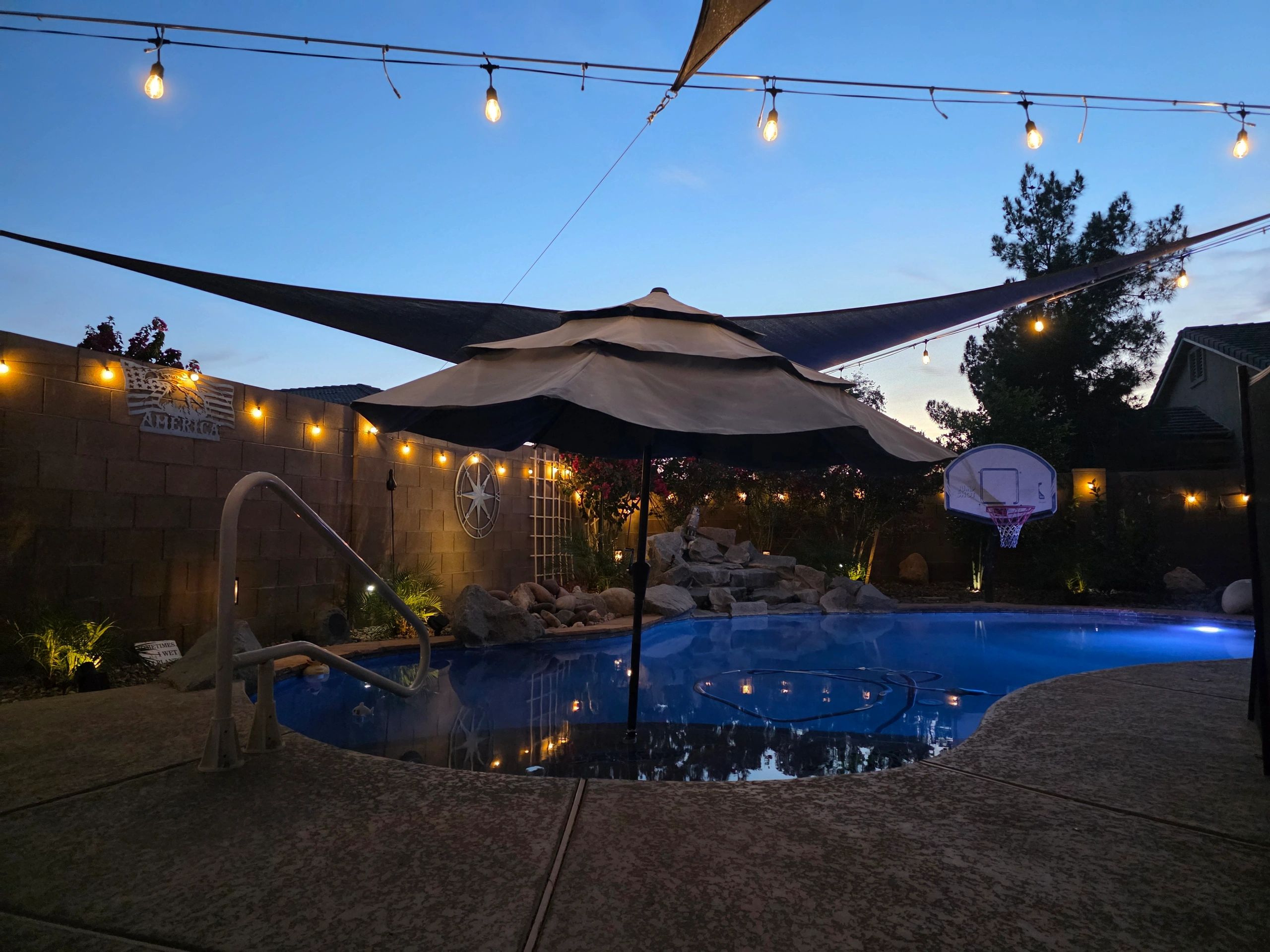 Evening backyard pool scene with umbrella, string lights, and basketball hoop.