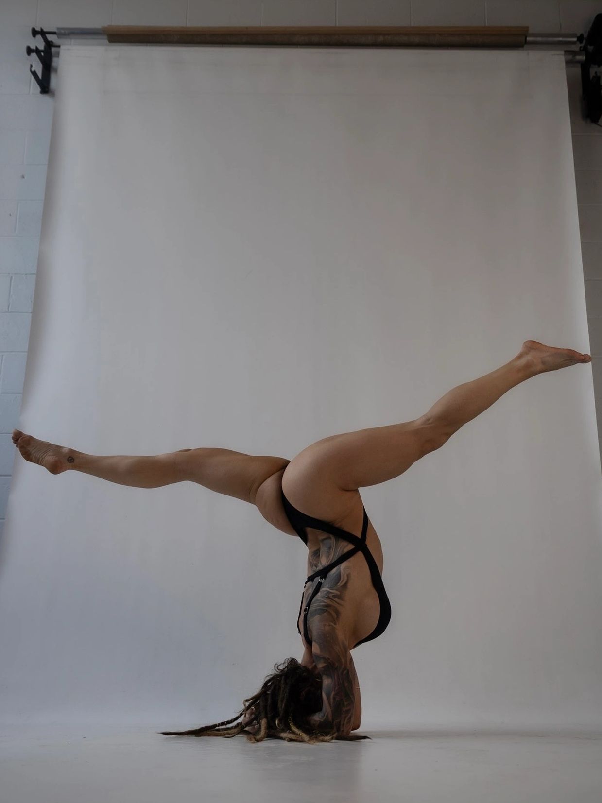 Person performing a handstand split with tattoos and dreadlocks against a plain backdrop.