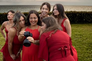 A group of six women in red dresses smiling at a camera screen outdoors.
