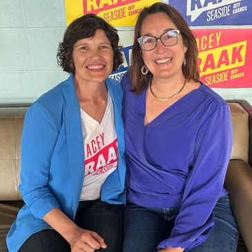 Two smiling women sitting together in front of colorful campaign signs.