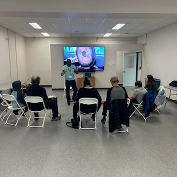 Group in a classroom learning with a presenter and gong on screen.