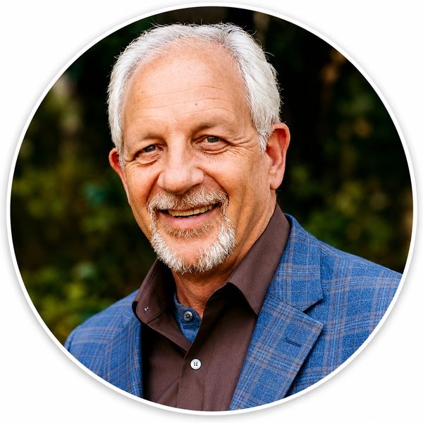 Smiling elderly man in a blue plaid blazer and brown shirt outdoors.