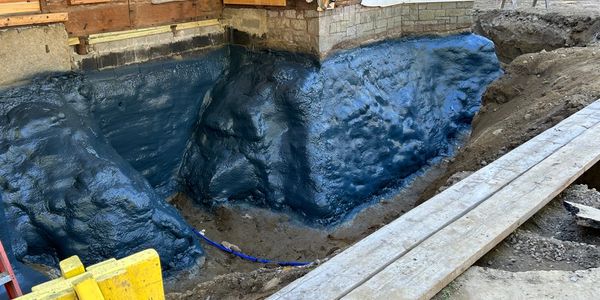 Excavated foundation area coated with a dark waterproofing material near a wooden structure.