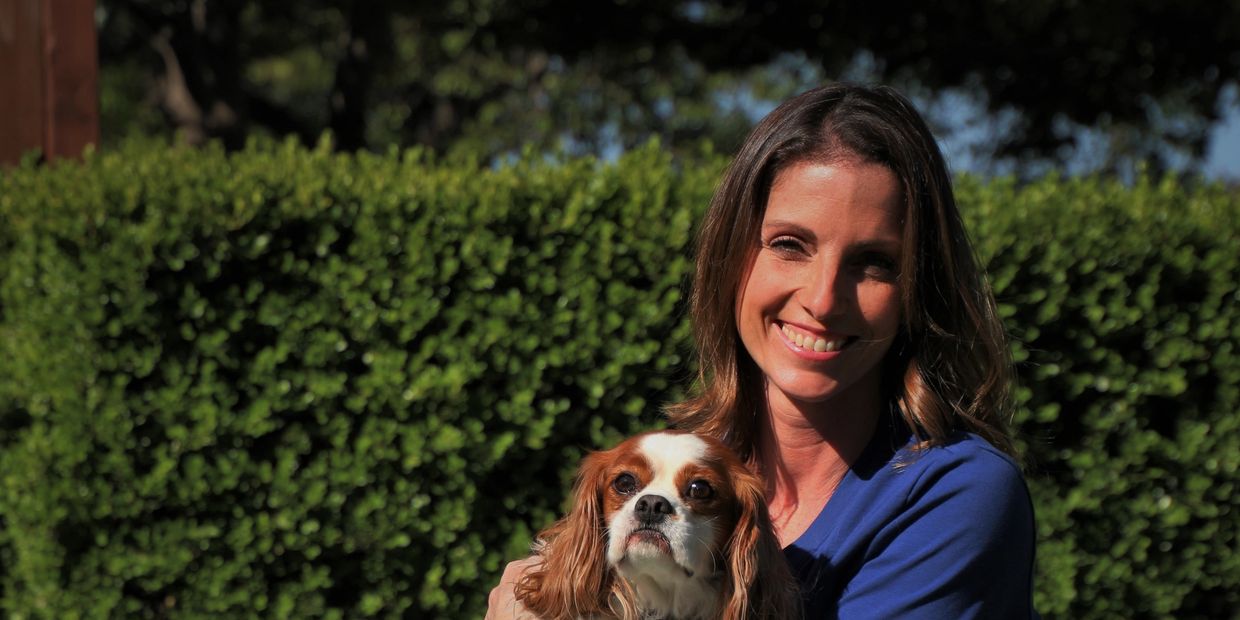 A smiling woman holds a small dog outdoors with greenery in the background.