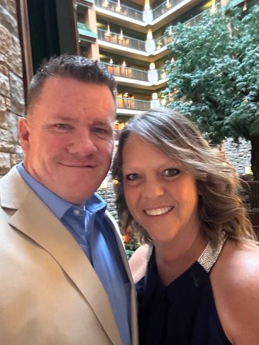 Couple smiling together indoors with a tree and balconies behind them.