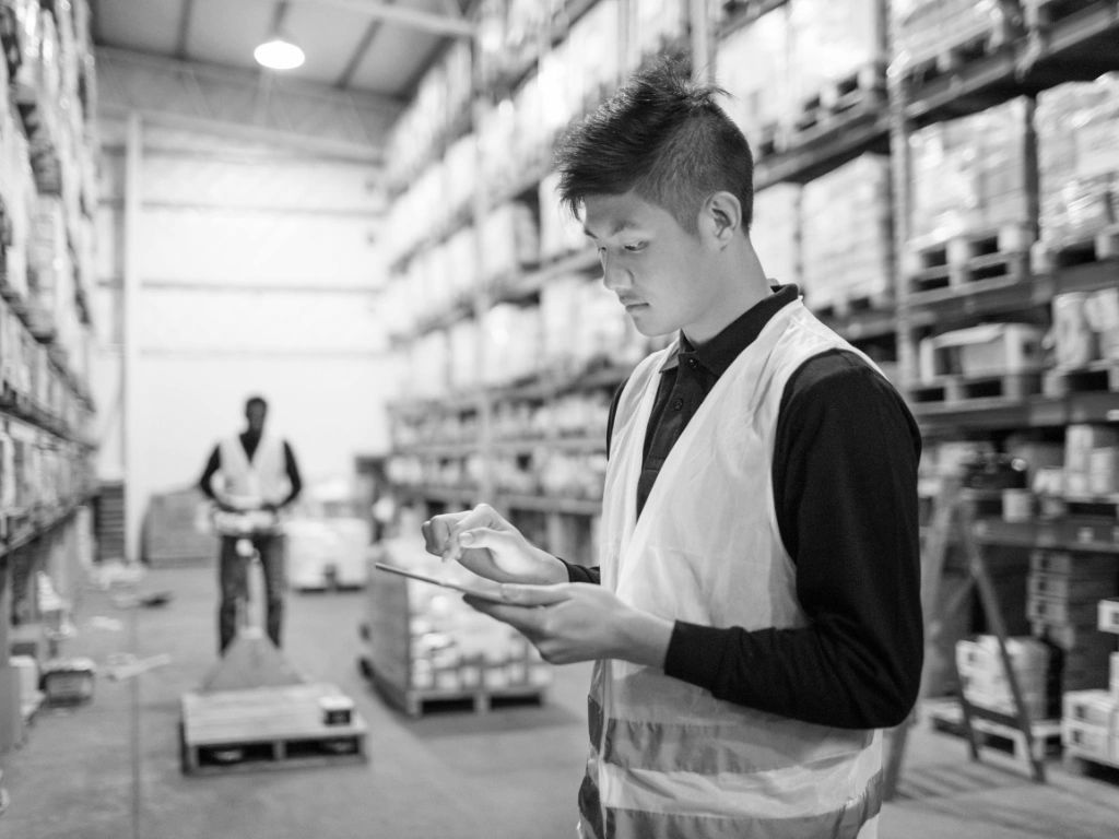 A warehouseman is standing in the warehouse with a tablet and behind him another warehouseman.
