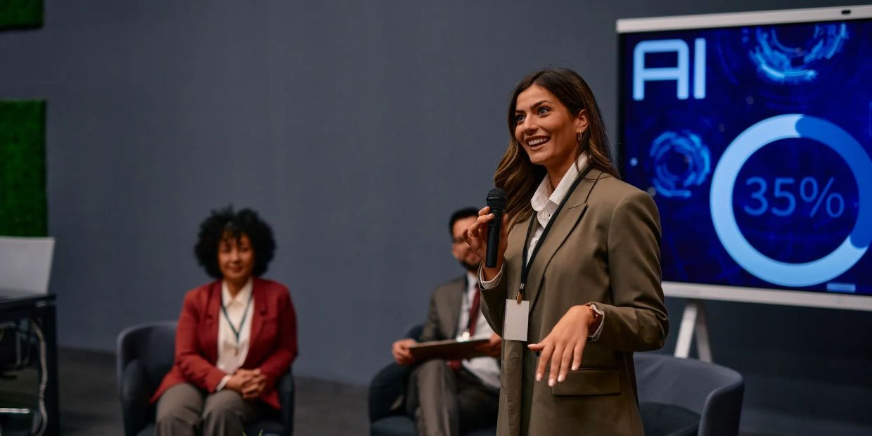 A woman speaks confidently at a tech conference with AI data displayed behind her.