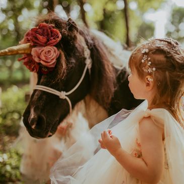 A girl in a dress looking at a unicorn pony adorned with flowers.