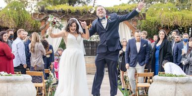Bride and groom joyfully jump together at their outdoor wedding ceremony.