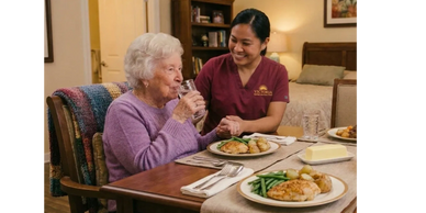 Elderly woman drinking water with a caregiver smiling beside her at a dining table.