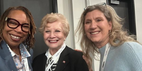 Three women smiling, wearing conference badges and lanyards indoors.
