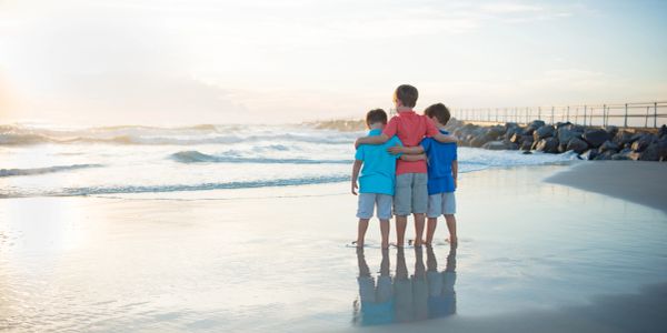 Three boys embrace while standing on a beach at sunrise.