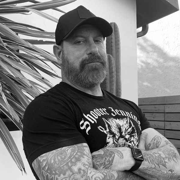 Bearded man with tattoos, wearing a cap and Shooter Jennings t-shirt, poses confidently outdoors.