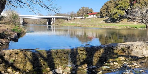 Calm river with a stone wall and bridge in the background on a clear day.